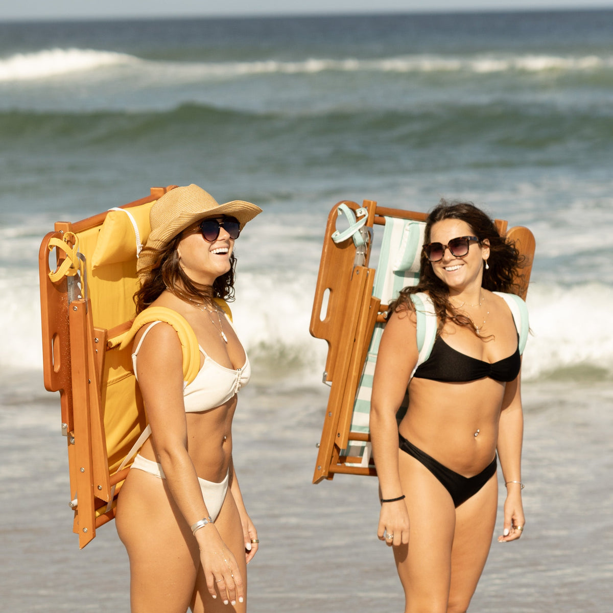 Two women carrying backpack beach chairs by the shore
