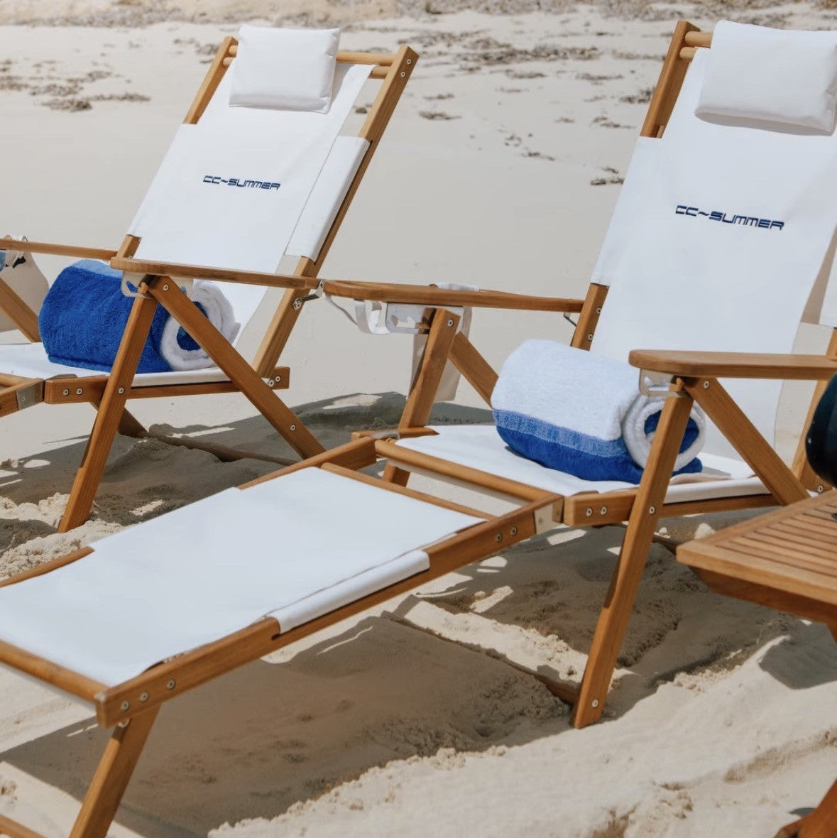 Teak beach lounge chairs in white that are personalized with an embroidered name, on a white sandy beach.