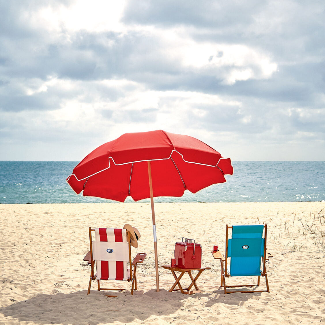 Red beach umbrella with two chairs and a small table on a sandy beach.