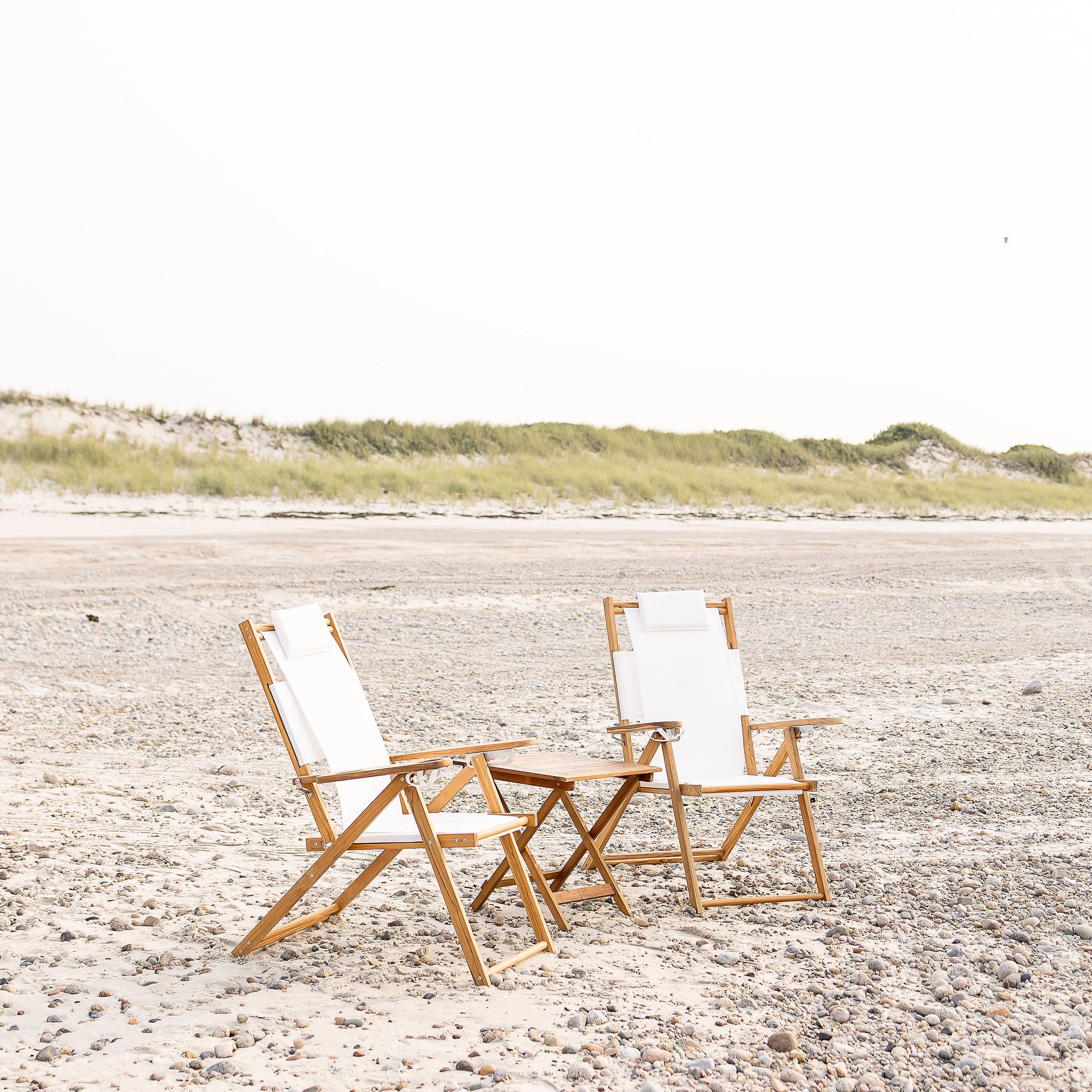 Two wooden beach chairs with white cushions on a sandy Nantucket beach.
