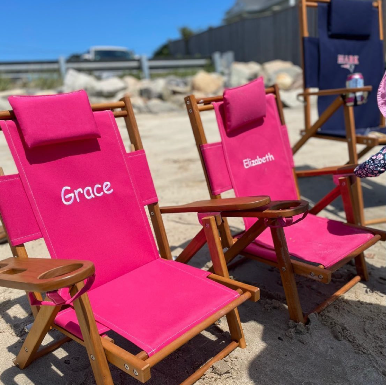 Two pink kid's beach chairs with names 'Grace' and 'Elizabeth' embroidered on a sandy beach.