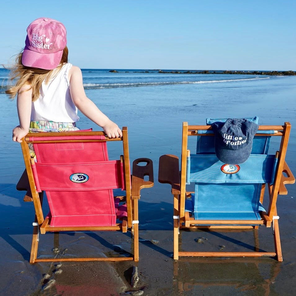 Coastal gender reveal featuring child with pink cap standing next to pink and baby blue custom child sized beach chairs on a sandy beach.