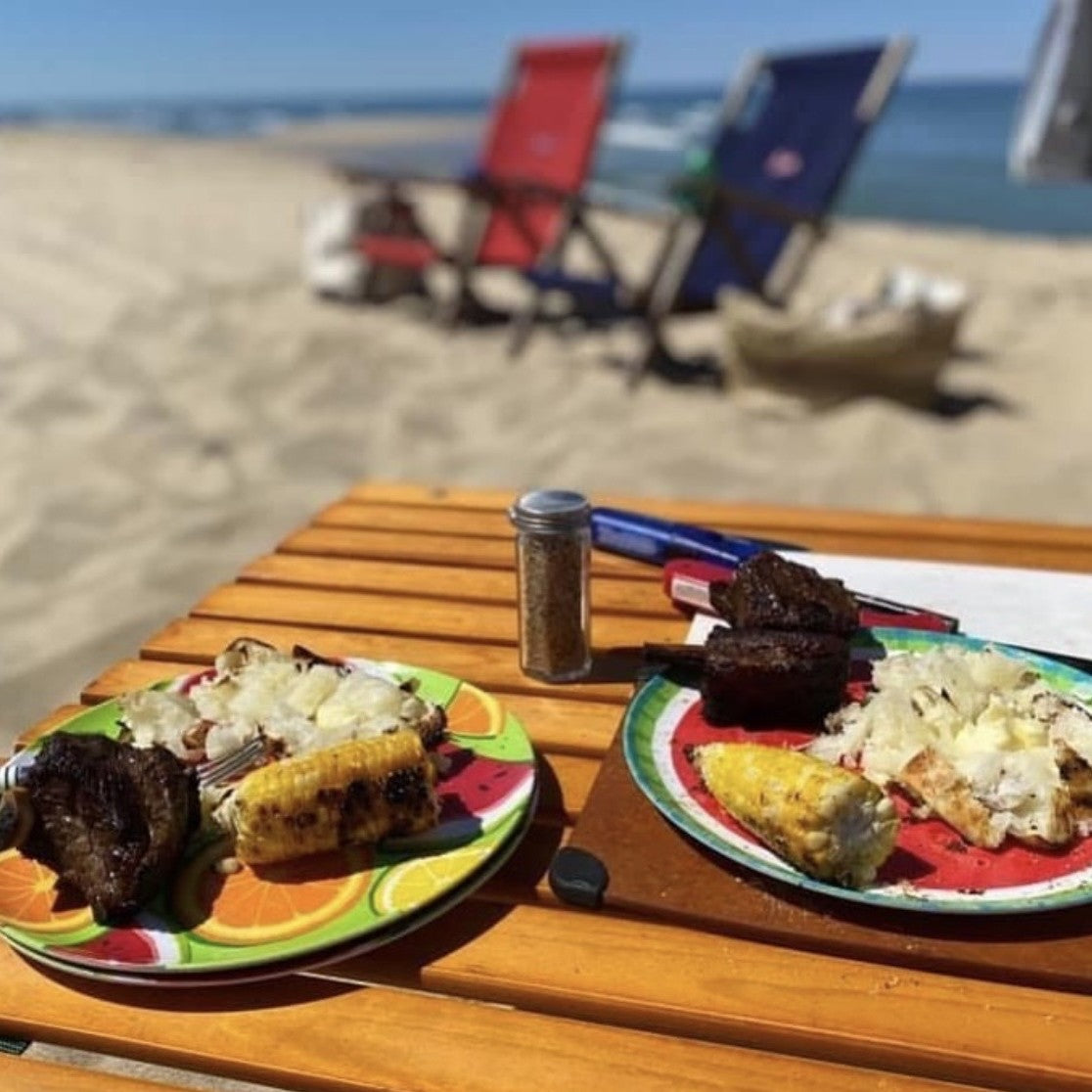 Two plates of food on a wooden table at the beach with chairs and sand in the background.