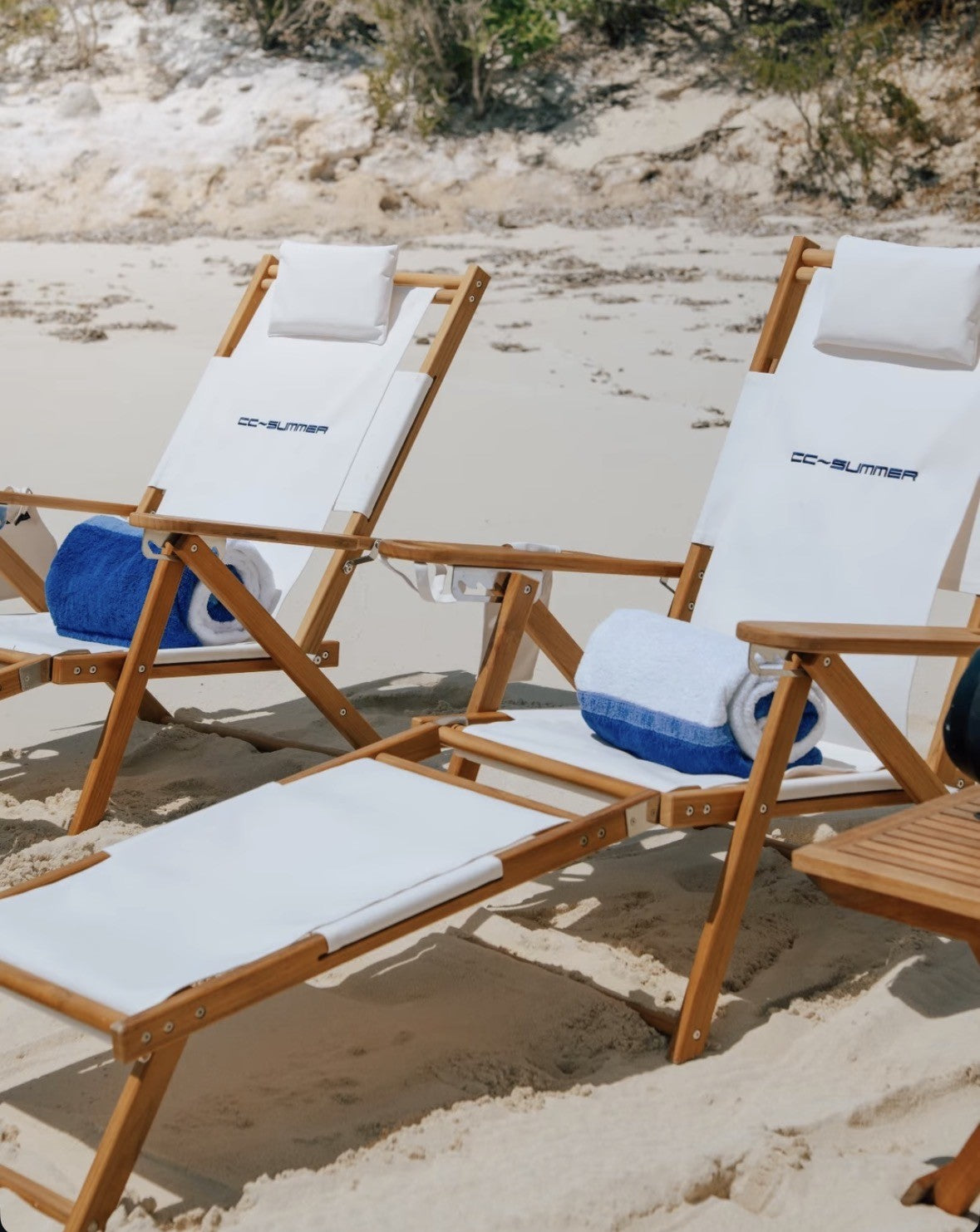 Teak beach lounge chairs in white that are personalized with an embroidered name, on a white sandy beach.