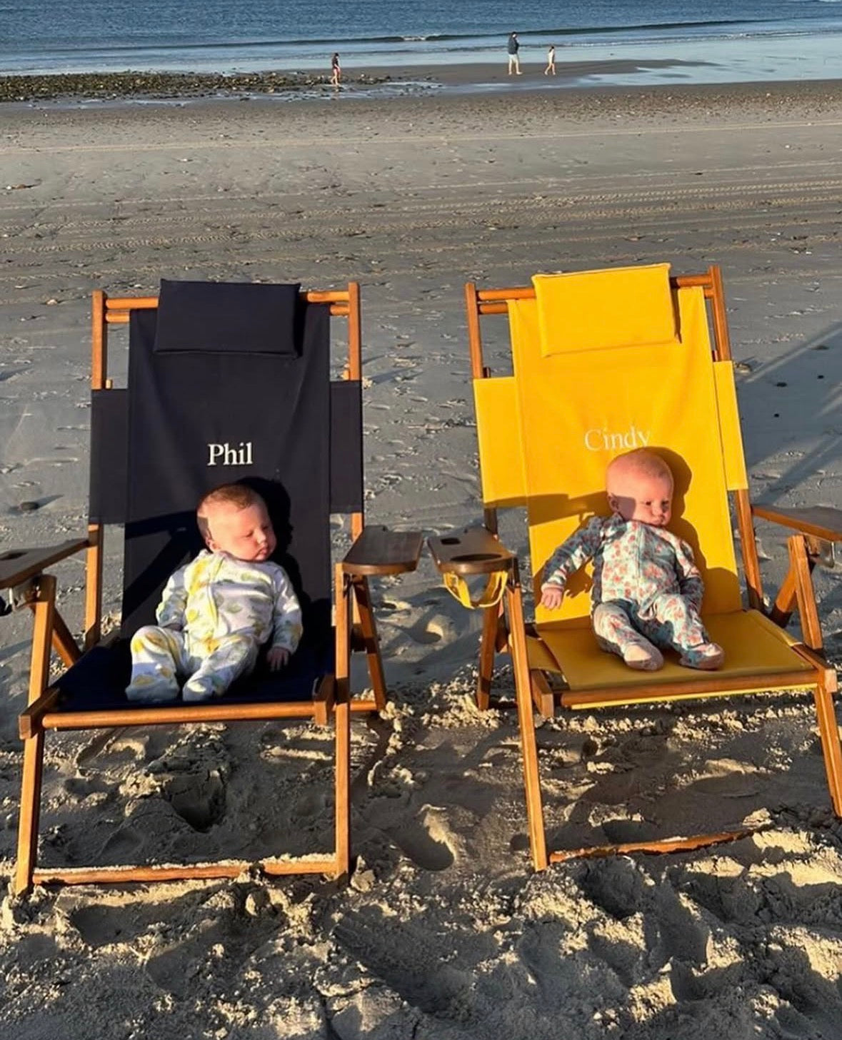 Two babies in wooden beach chairs named 'Phil' and 'Cindy' on a sandy beach.