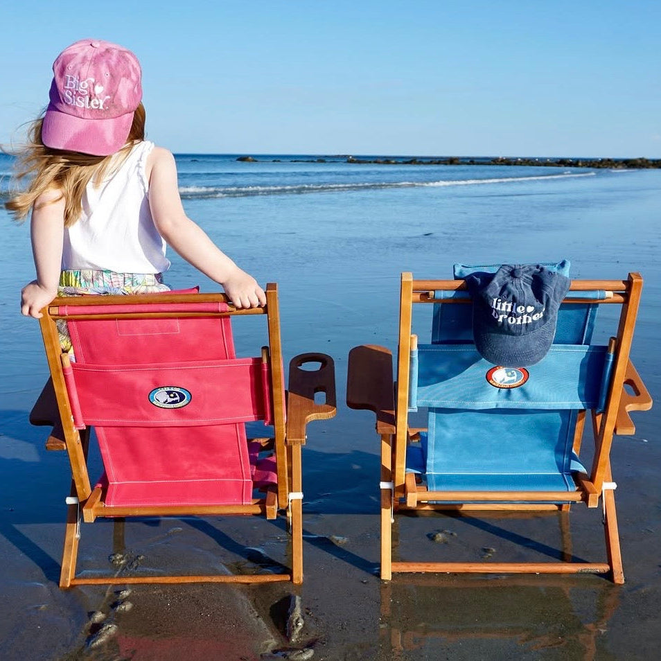 Child with pink cap standing next to children's sized beach chairs on a sandy beach for a coastal gender reveal.