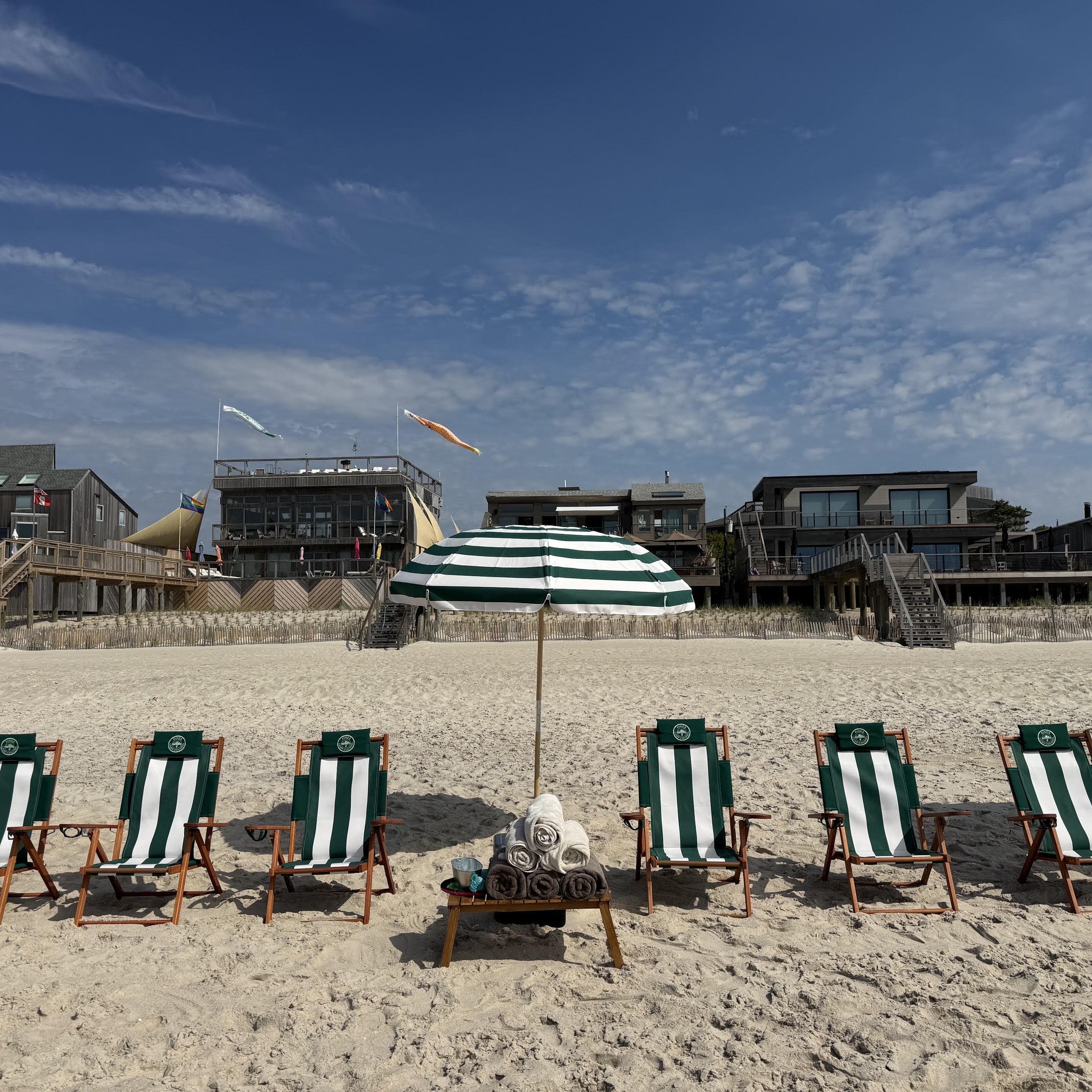 Fire Island beach scene with green and white striped chairs and umbrella on a sandy beach with buildings in the background.