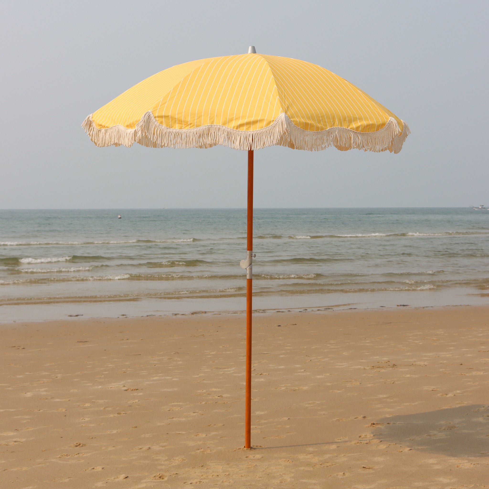 Yellow wood poled beach umbrella with white fringe on a sandy beach with ocean in the background